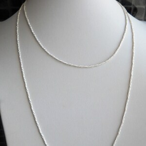 May include: Two silver-colored necklaces displayed on a white mannequin. The necklaces are delicate, with a twisted rope design. The longer necklace hangs down, while the shorter one rests higher on the mannequin's neck. The background is neutral.