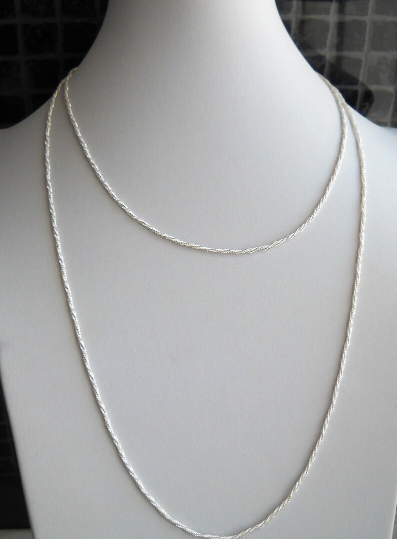 May include: Two silver-colored necklaces displayed on a white mannequin. The necklaces are delicate, with a twisted rope design. The longer necklace hangs down, while the shorter one rests higher on the mannequin's neck. The background is neutral.