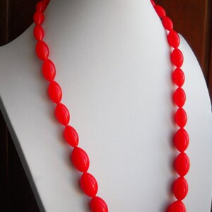 Puede incluir: Un collar rojo vibrante compuesto por cuentas de forma ovalada. Las cuentas están ensartadas, creando un bucle continuo. El collar se exhibe en un maniquí blanco, mostrando su diseño y color. Las cuentas tienen un acabado brillante.