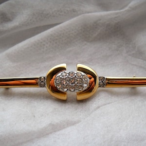 Può includere: Un fermaglio in oro e argento con un design ovale centrale incastonato con strass trasparenti. Il fermaglio ha una lunga barra sottile con strass più piccoli su ciascuna estremità.