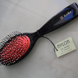 Pode incluir: Uma escova de cabelo preta com cerdas vermelhas e um cabo preto. O cabo tem o texto "GéBé" em letras douradas. A escova tem uma etiqueta anexada que diz "NYLON POUR PÉNÉTRER ET COIFFER MADE IN FRANCE".