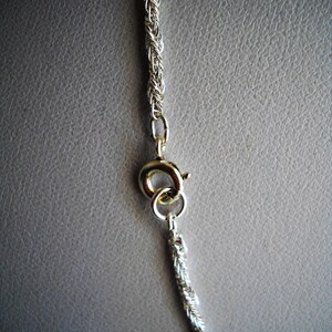 May include: Close-up of a silver-colored rope chain necklace with a gold-colored clasp. The chain is made of tightly woven strands, and the clasp is a simple, circular design. The background is a neutral gray.