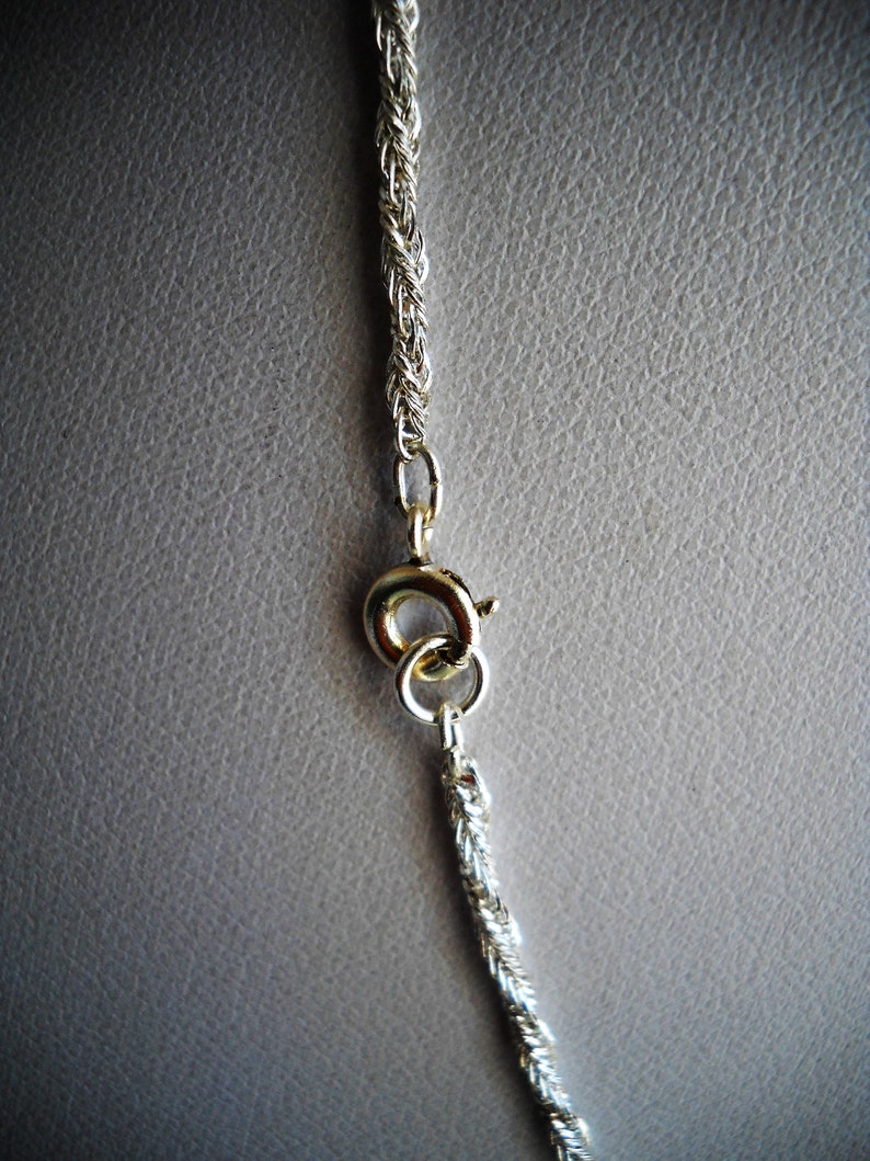 May include: Close-up of a silver-colored rope chain necklace with a gold-colored clasp. The chain is made of tightly woven strands, and the clasp is a simple, circular design. The background is a neutral gray.