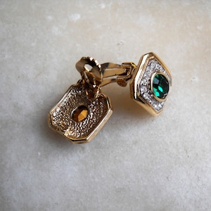 Pode incluir: Um par de brincos de pressão dourados com uma gema verde cercada por strass transparentes.