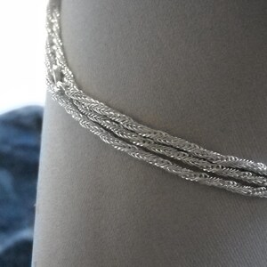 May include: A silver-colored bracelet with a braided rope design. The bracelet is composed of three strands of tightly woven metal, creating a textured appearance. The jewelry is displayed against a neutral background.