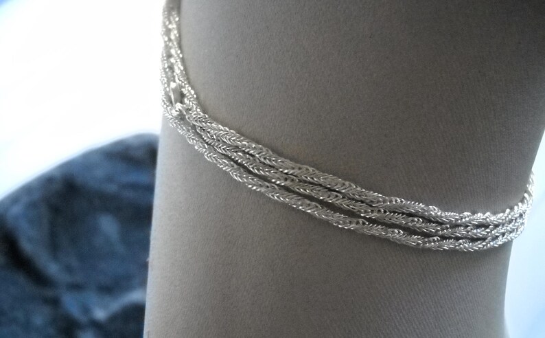 May include: A silver-colored bracelet with a braided rope design. The bracelet is composed of three strands of tightly woven metal, creating a textured appearance. The jewelry is displayed against a neutral background.