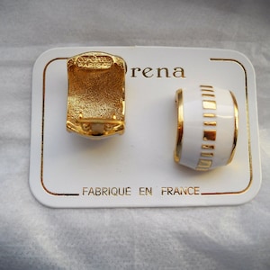 Può includere: Un paio di orecchini a cerchio dorati e smaltati bianchi. Un orecchino è dorato, l'altro è bianco con dettagli dorati. Gli orecchini sono su una carta bianca con la scritta "Fabriqué en France".