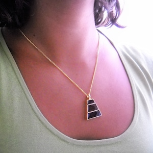 Pode incluir: Um colar de corrente de ouro com um pingente preto em forma de pirâmide. O pingente tem três faixas horizontais pretas.