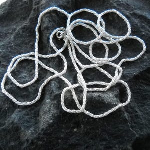 May include: A silver-colored necklace with a textured, rope-like design. The necklace is lying on a dark, textured surface. The clasp is visible, indicating the necklace is ready to wear.