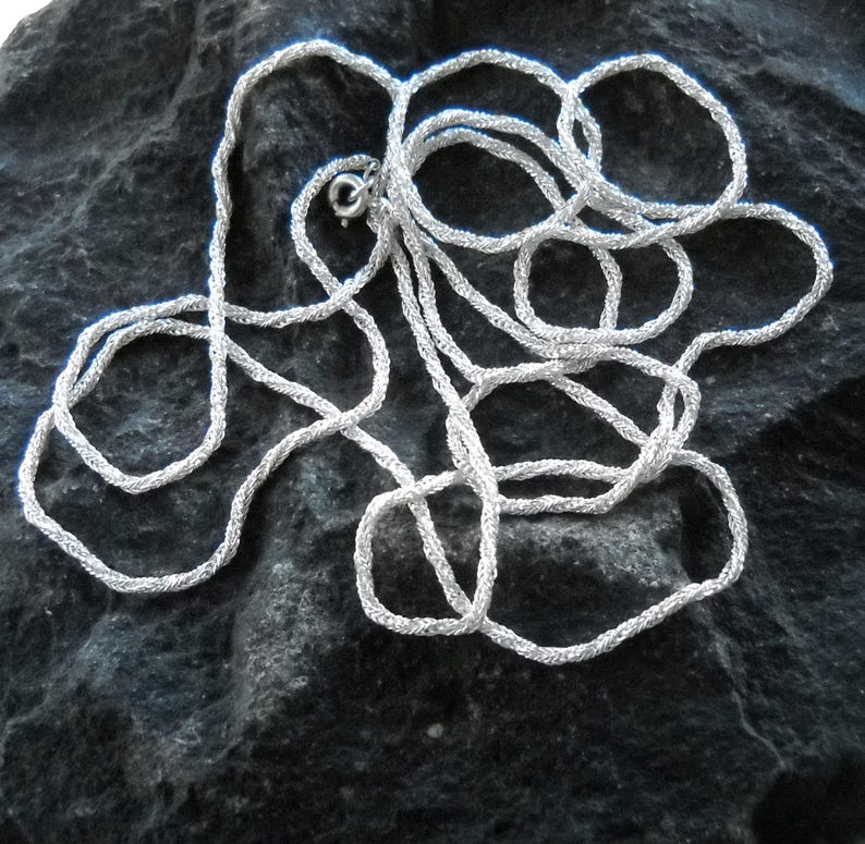 May include: A silver-colored necklace with a textured, rope-like design. The necklace is lying on a dark, textured surface. The clasp is visible, indicating the necklace is ready to wear.