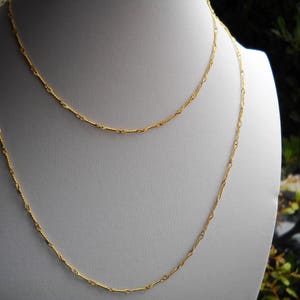 May include: Two gold chains, one thicker than the other, are layered on a white mannequin bust. The chains are made of a delicate, twisted design.