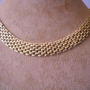 May include: A gold chain necklace with a woven, box-style design. The necklace is on a brown velvet background.
