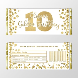 Puede incluir: Envoltorio de barra de chocolate dorada y blanca con un diseño de purpurina dorada. El envoltorio dice "Olivia's Golden Birthday" y tiene un número 10 dorado. La parte posterior del envoltorio tiene una etiqueta de información nutricional con el texto "¡Gracias por celebrar conmigo!" y "Happy Day".
