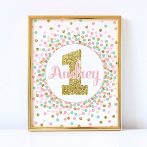 May include: Gold glitter number one with the name Audrey in pink script on a white background with pink and green confetti.