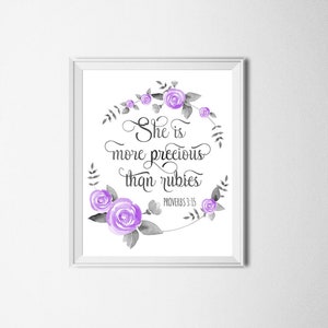 Puede incluir: Una impresión enmarcada en blanco con una corona floral púrpura y gris que rodea el texto "Ella es más preciosa que las rubíes" con la referencia bíblica "Proverbios 3:15" debajo.