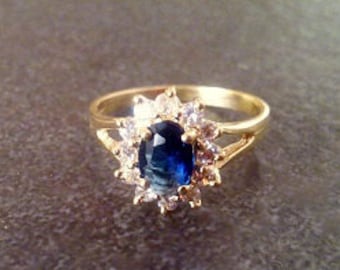 Könnte beinhalten: Ein goldener Ring mit einem blauen ovalen Edelstein, der von kleineren klaren Edelsteinen umgeben ist.