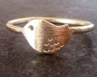 May include: A gold-colored ring featuring a bird design. The bird has a circular eye and a textured body with small, circular cutouts. The ring band is smooth and simple, and the ring is set against a dark background.