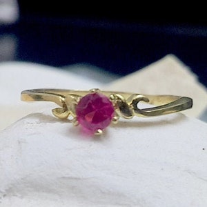 Könnte beinhalten: Ein goldener Ring mit einem rosafarbenen Edelstein in der Mitte. Der Ring hat ein zartes Design mit zwei geschwungenen Linien auf jeder Seite des Steins.