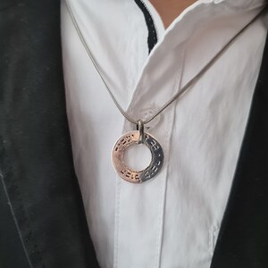 Puede incluir: Un collar de cadena de plata con un colgante redondo con texto en hebreo. El colgante se lleva sobre una camisa blanca de cuello y una chaqueta negra.