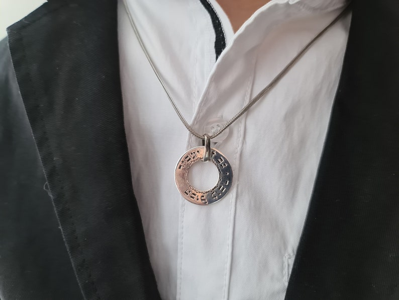 Puede incluir: Un collar de cadena de plata con un colgante redondo con texto en hebreo. El colgante se lleva sobre una camisa blanca de cuello y una chaqueta negra.