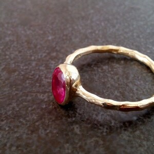 K&ouml;nnte beinhalten: Ein goldener Ring mit einem leuchtend pinkfarbenen ovalen Edelstein, der in einer Zargenfassung sitzt. Die Ringschiene ist texturiert und hat eine geh&auml;mmerte Oberfl&auml;che.