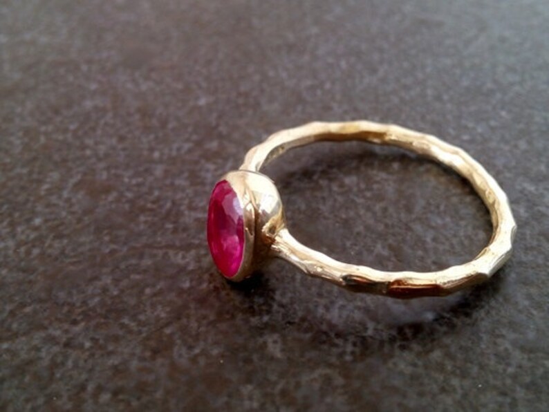 K&ouml;nnte beinhalten: Ein goldener Ring mit einem leuchtend pinkfarbenen ovalen Edelstein, der in einer Zargenfassung sitzt. Die Ringschiene ist texturiert und hat eine geh&auml;mmerte Oberfl&auml;che.