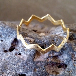 Può includere: Un anello d'oro con un design a zig-zag irregolare. L'anello è in metallo liscio e lucido e ha uno stile semplice e moderno.