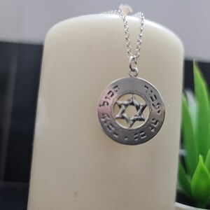 Puede incluir: Un collar de plata con un colgante redondo que presenta una estrella de David y texto en hebreo. El colgante est&aacute; en una cadena de plata.