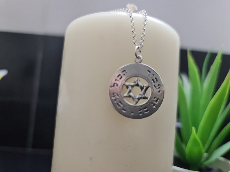 Puede incluir: Un collar de plata con un colgante redondo que presenta una estrella de David y texto en hebreo. El colgante est&aacute; en una cadena de plata.