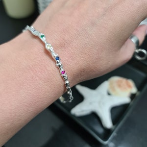 Puede incluir: Un brazalete de plata con un diseño trenzado y piedras preciosas de colores. Las piedras preciosas están dispuestas en una fila a lo largo del brazalete y son rojas, azules, verdes y amarillas.