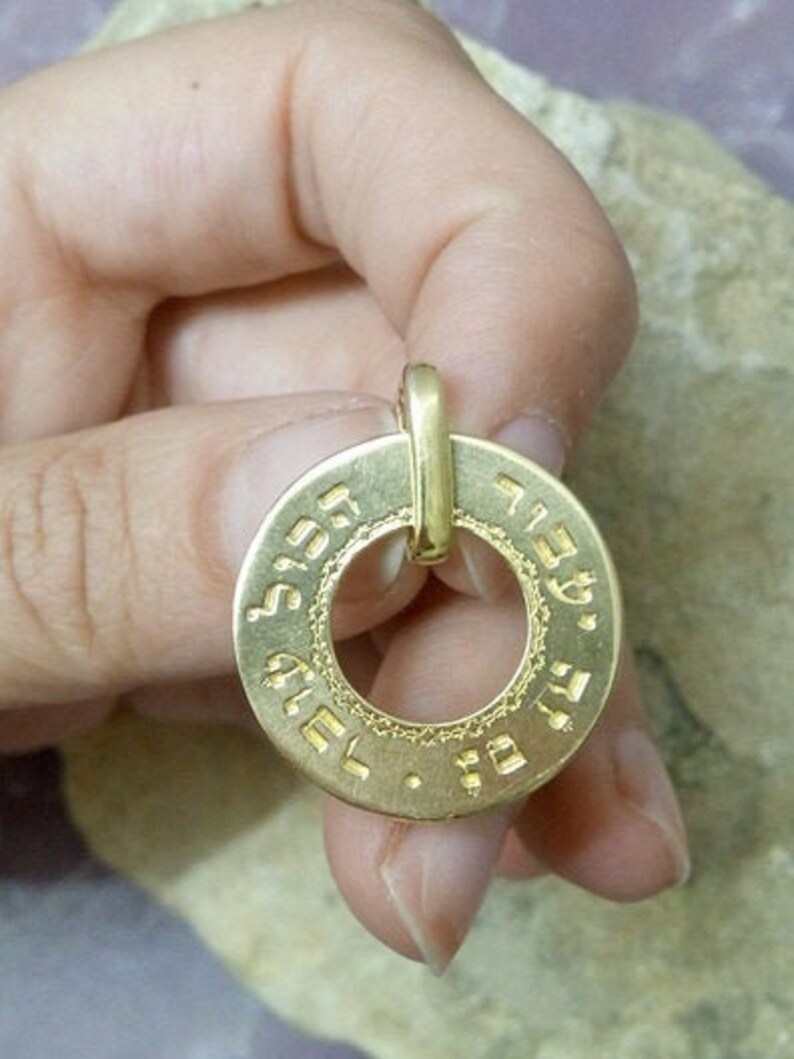 Puede incluir: Pendiente de oro con forma redonda y escritura hebrea alrededor del borde. El pendiente tiene un agujero en el centro para una cadena.