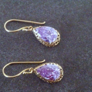 May include: A pair of gold earrings with a teardrop-shaped purple gemstone set in a gold filigree frame.