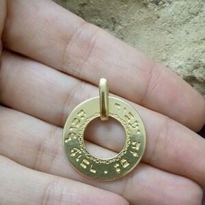 Puede incluir: Un colgante de oro con un dise&ntilde;o circular que presenta texto hebreo alrededor del borde. El colgante tiene un bucle en la parte superior para sujetarlo a una cadena.