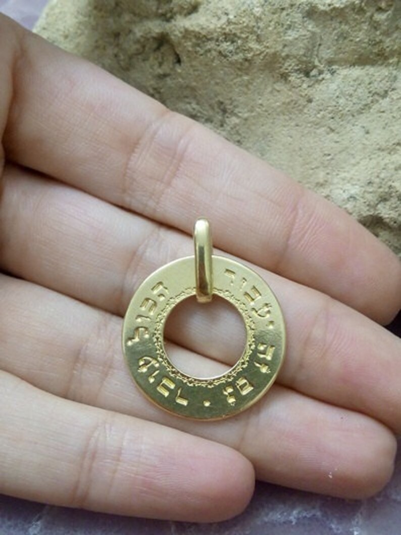 Puede incluir: Un colgante de oro con un dise&ntilde;o circular que presenta texto hebreo alrededor del borde. El colgante tiene un bucle en la parte superior para sujetarlo a una cadena.