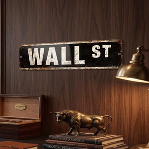 Op de afbeelding: Een rechthoekig metalen straatnaambord met de tekst Wall St in een verweerde zwart-witte afwerking aan een donkere houten muur, boven een bureau met een bronzen stierenbeeld, een houten doos, een notitieboek en financiële tijdschriften.