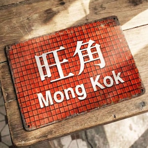 May include: A rectangular red metal sign with white text that reads "旺角 Mong Kok". The sign has a grid pattern and is placed on a wooden table. The sign is slightly weathered, giving it a vintage look.