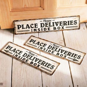 Puede incluir: Tres letreros rectangulares de metal con el texto "Please PLACE DELIVERIES INSIDE BOX" en letras negras. Los letreros tienen un aspecto envejecido y vintage con fondo blanco y borde marrón. Un letrero tiene una flecha.