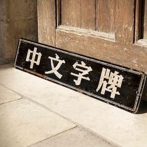 Könnte beinhalten: Ein rechteckiges, verwittertes schwarzes Schild mit weißen chinesischen Schriftzeichen, das an einer Holztür lehnt. Das Schild trägt die Aufschrift "Chinesisches Schriftzeichen". Das Schild steht auf einer hellen Steinoberfläche.