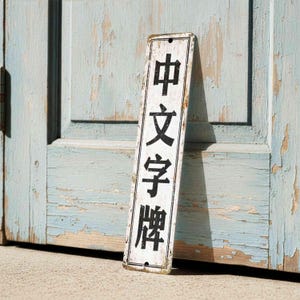 Könnte beinhalten: Ein rechteckiges Schild im Vintage-Stil mit schwarzen chinesischen Schriftzeichen auf weißem Grund. Das Schild lehnt an einer verwitterten, hellblauen Tür. Die Kanten des Schildes weisen Gebrauchsspuren auf, was ihm ein rustikales Aussehen verleiht.