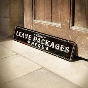 May include: A rectangular black and white sign with the text "LEAVE PACKAGES HERE" in a distressed font. The sign is leaning against a wooden door and a stone wall. The sign has a vintage, rustic aesthetic.