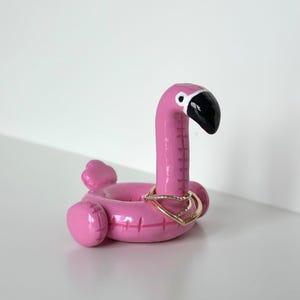 Może przedstawiać: Różowy uchwyt na pierścionki w kształcie flaminga z czarnym dziobem i okiem. Uchwyt na pierścionki ma złoty pierścionek. Flaming stoi na białej powierzchni.