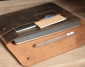 May include: A brown leather laptop sleeve with a flap closure and a pocket for a pen or stylus. The sleeve is open and a laptop, notebook, and phone are inside.