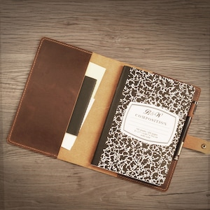 Puede incluir: Una cubierta de cuaderno de cuero marrón con un cuaderno de composición en blanco y negro en el interior. El cuaderno tiene el texto "B&W Composition" en la portada. La cubierta tiene un portaplumas y una correa con un botón.