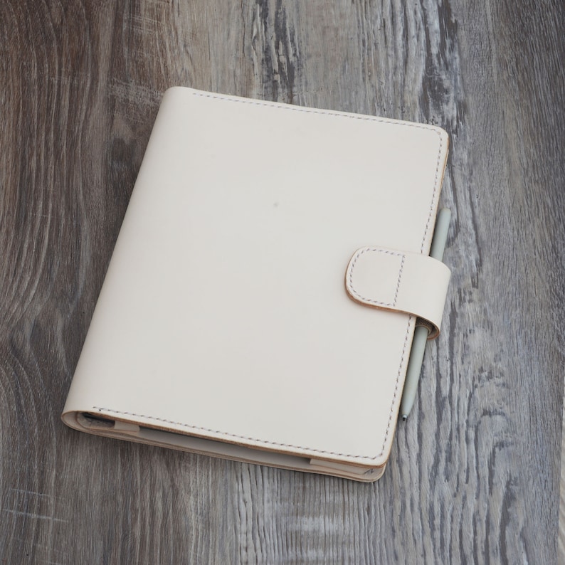 May include: Cream-colored leather journal with a matching closure strap and a gray stylus. The journal is closed and has a stitched border. The journal is on a wooden surface.