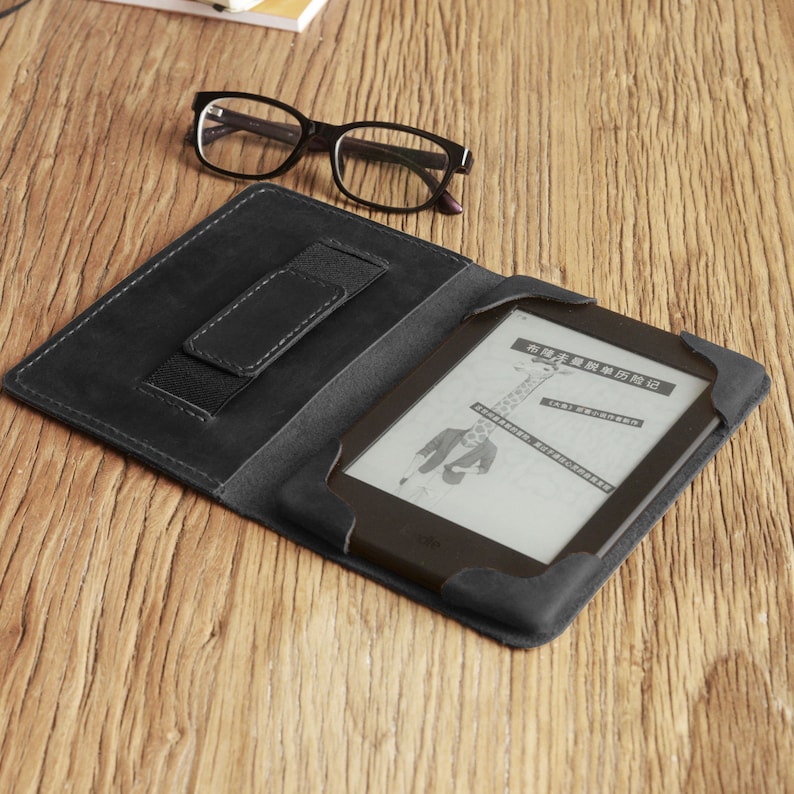 Puede incluir: Una funda de cuero negro para lector electr&oacute;nico con un bolsillo integrado para una tarjeta de cr&eacute;dito o identificaci&oacute;n. La funda est&aacute; abierta y un lector electr&oacute;nico est&aacute; dentro. La pantalla del lector electr&oacute;nico muestra una p&aacute;gina de texto en chino.