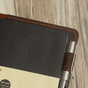 May include: A brown leather notebook with a pen holder and a soft cover. The notebook has a stitched edge and the text "soft cover" is printed on the front.