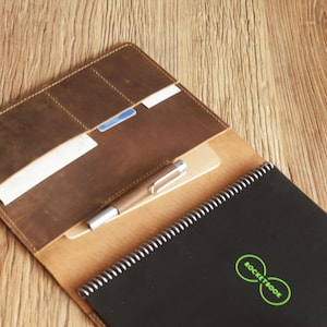 K&ouml;nnte beinhalten: Ein braunes Leder-Notizbuchcover mit einem Stift und Karten im Inneren. Das Notizbuch hat einen schwarzen Einband mit einem gr&uuml;nen Rocketbook-Logo.