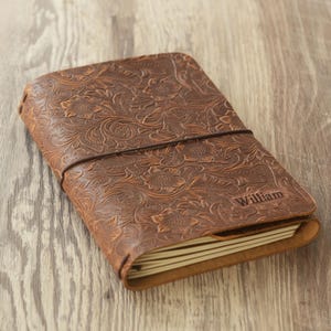 May include: A brown leather journal with an embossed floral pattern. The journal is closed with an elastic band and has the name "William" embossed on the cover. The pages are visible along the edge.