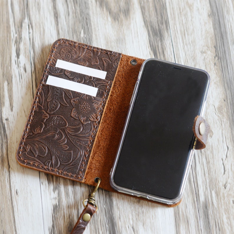 K&ouml;nnte beinhalten: Eine braune Leder Handyh&uuml;lle mit einem gepr&auml;gten Blumenmuster. Die H&uuml;lle hat eine Klappe, die &uuml;ber das Handy geklappt wird, und ein Fach f&uuml;r Karten. Die H&uuml;lle hat einen Handgelenksriemen.