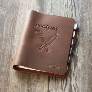 Puede incluir: Una carpeta de recetas de cuero marrón con la palabra recipes en relieve y un gráfico de un batidor y un cuchillo cruzados. El nombre Maddy está grabado en la esquina inferior derecha. La carpeta incluye varias pestañas divisorias de colores.